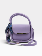 A lavender purple top-handle crossbody bag with a matching shoulder strap, a gold-tone metal bar on the front, and a blue patterned silk scarf tied to the side, shown on a light gray background.