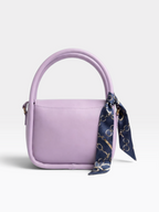 A lavender purple leather handbag with a top handle, adorned with a navy blue silk scarf featuring a gold chain pattern tied to the side, set against a plain light gray background.