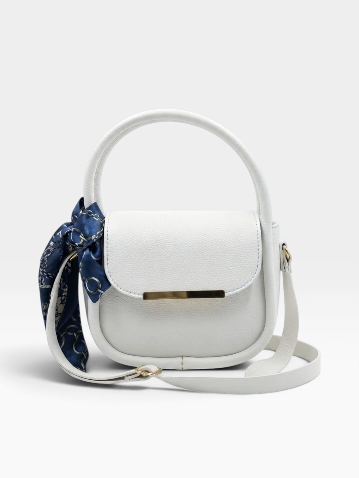 A white pebbled leather top-handle shoulder bag with a matching adjustable shoulder strap, a gold-tone horizontal bar accent on the front, and a navy blue patterned silk scarf tied to the side, shown against a light gray studio background.