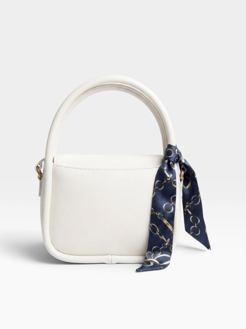 A structured white leather handbag with a top handle and a navy blue silk scarf featuring a gold chain-link pattern tied to the side, shown on a light gray background.
