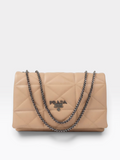 Prada quilted flap shoulder bag in beige leather with a silver-tone chain strap and a silver-tone Prada logo plaque on the front, photographed on a light gray background.