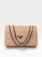 Prada quilted flap shoulder bag in beige leather with a silver-tone chain strap and a silver-tone Prada logo plaque on the front, photographed on a light gray background.