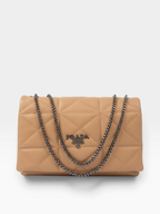 A Prada quilted shoulder bag in tan leather with a silver-tone chain strap and a silver-tone metal logo plaque on the front, set against a light gray background.
